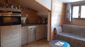 A compact kitchen with a Siemens oven, a dishwasher, and a coffee maker. There's a wooden ceiling, a window with curtains, and a sofa in the living area.