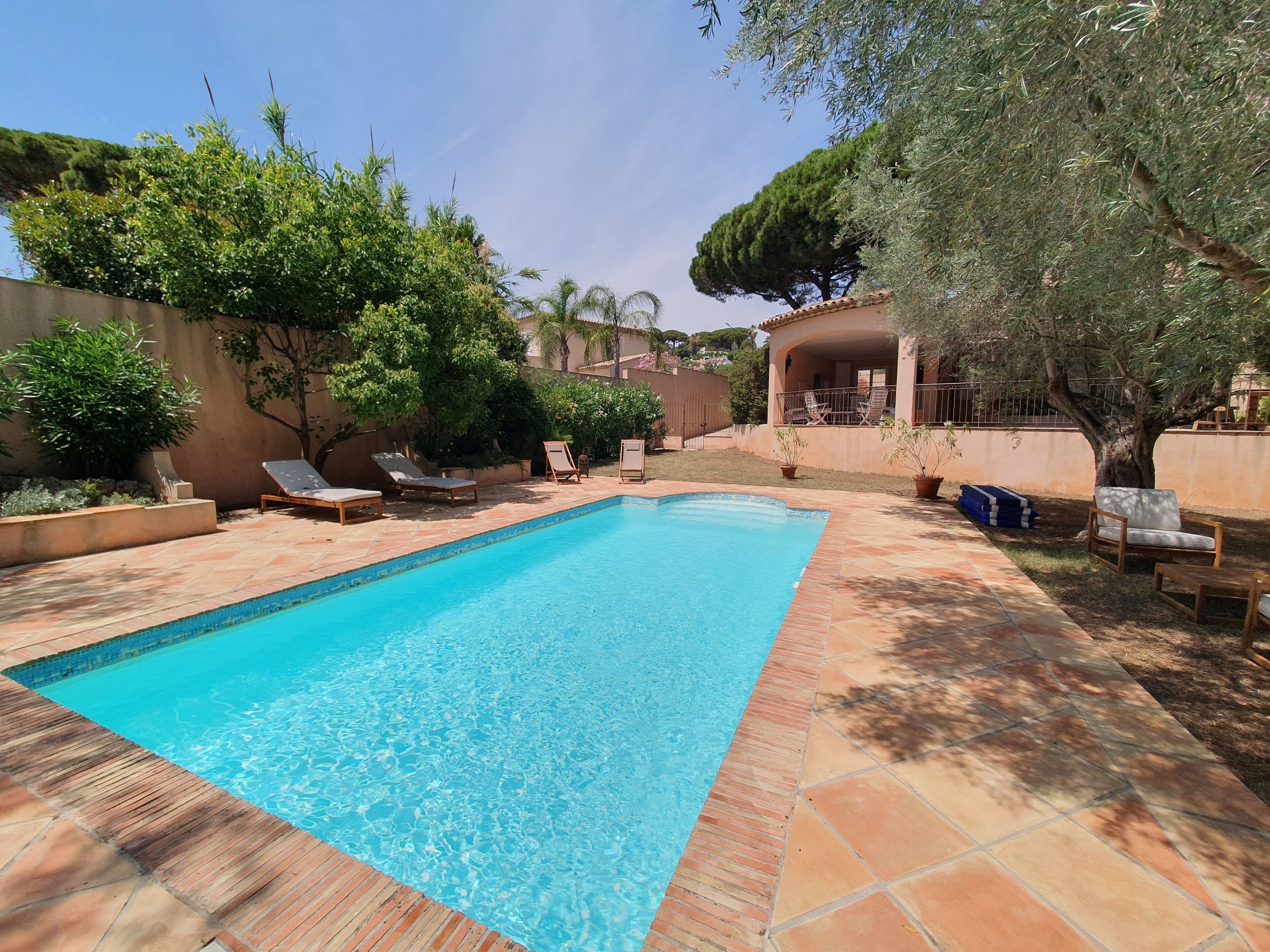 A rectangular swimming pool with clear blue water, surrounded by a tiled deck. There are lounge chairs and a covered seating area with a pergola.