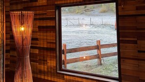 Una habitación con paredes de madera, un ventanal con vistas a un área cercada y una lámpara alta y cilíndrica.
