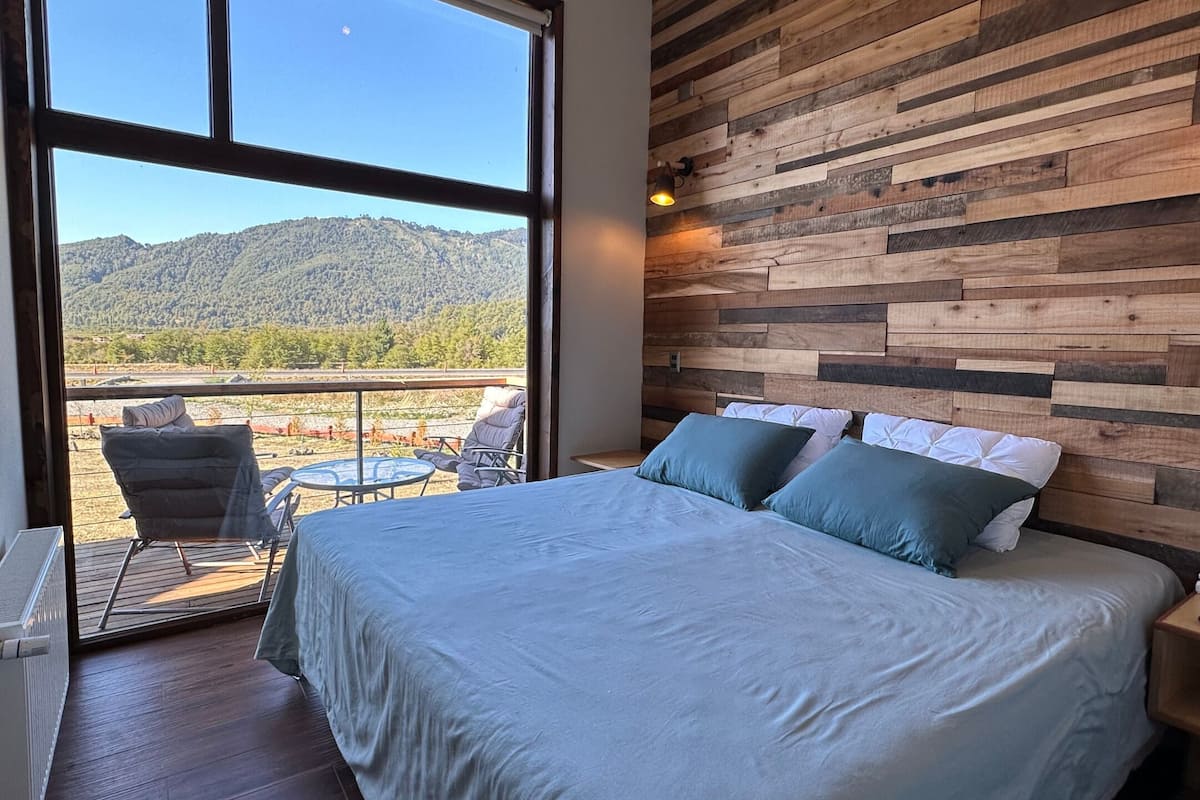 Ein Schlafzimmer mit einem großen Bett, Blick auf eine gebirgige Landschaft durch ein Fenster und einer Holzwand.