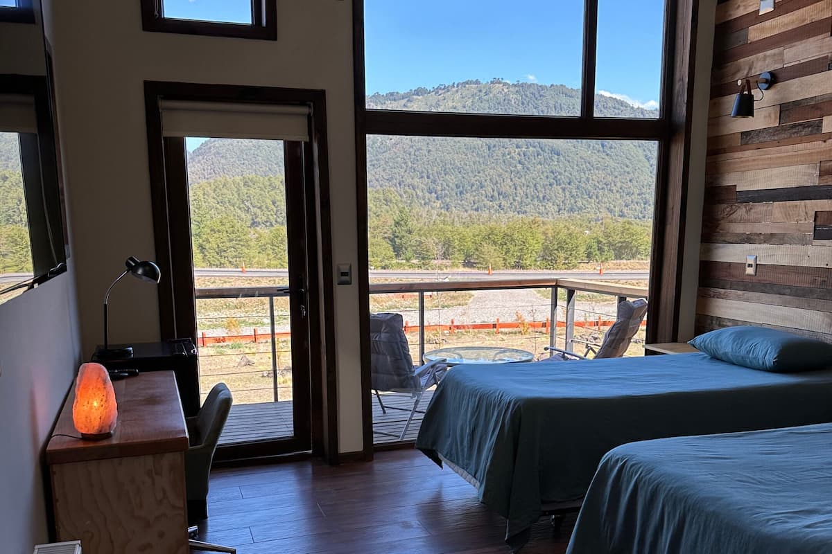 Ein Schlafzimmer mit zwei Betten, einem Schreibtisch mit Lampe und Blick auf eine gebirgige Landschaft durch große Fenster.
