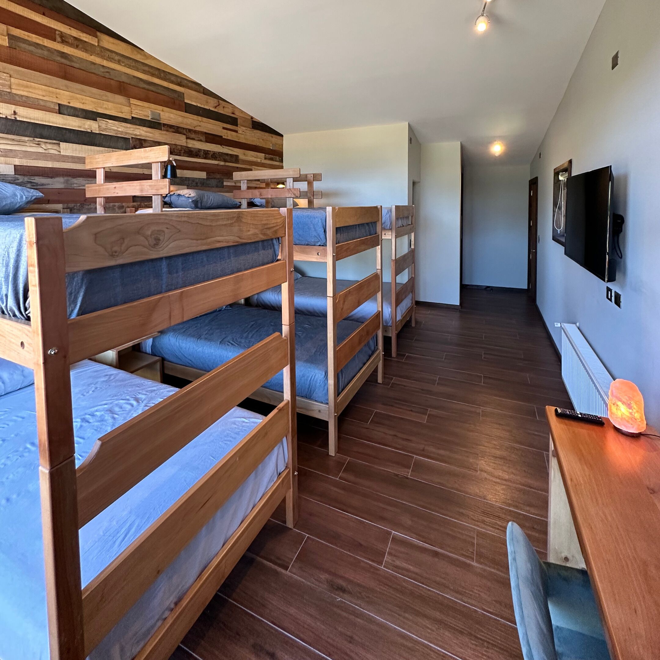 A dormitory room with bunk beds, a wooden floor, a desk with a lamp, and a TV mounted on the wall.