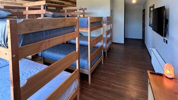 A dormitory room with bunk beds, a wooden floor, a desk with a lamp, and a TV mounted on the wall.