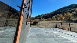 Vista desde el interior de un edificio hacia una terraza empedrada con barandilla metálica, cielo azul despejado y un paisaje montañoso con árboles al fondo.