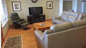 Uma sala de estar com sofá, poltrona, mesa de centro, televisão e quadros nas paredes.