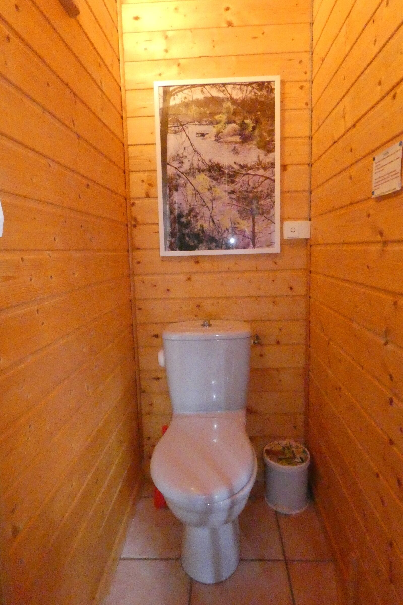 Une petite salle de bain aux murs lambrissés, équipée de toilettes, d’une poubelle et d’un tableau encadré.