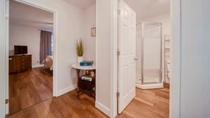 Una habitación con piso de madera, una puerta blanca, una mesita con una planta y una zona de ducha visible.