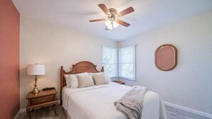 A bedroom with a bed, nightstand, lamp, ceiling fan, window, and wall mirror.
