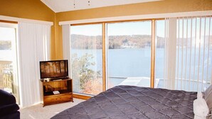 Un dormitorio con una cama grande, un televisor sobre un soporte de madera y vista a un lago a través de amplias ventanas.