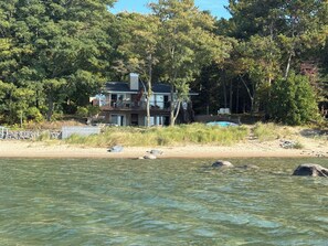 Una casa con balcón que da a una masa de agua, rodeada de árboles y una playa de arena.