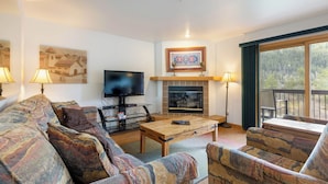 A living room with a sofa, a coffee table, a fireplace, a TV, and a balcony with a view of trees. - Panoramic Mountain Views/Hot Tub Access/Private Deck with Grill (Keystone)