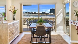A dining area with a table and chairs, a plant on the table, and a view of the beach and buildings through the glass doors. - Baybreeze-3bd, 2ba, sleep 8 (San Diego)