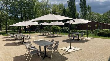 An outdoor patio area with tables and chairs, shaded by large umbrellas, set against a backdrop of trees and a building.
