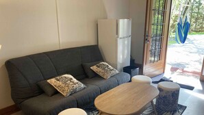 Una sala de estar moderna con un sofá gris, dos cojines a juego, una mesa de centro de madera y dos otomanas redondas. Un refrigerador blanco está contra la pared, y una alfombra estampada cubre el suelo.