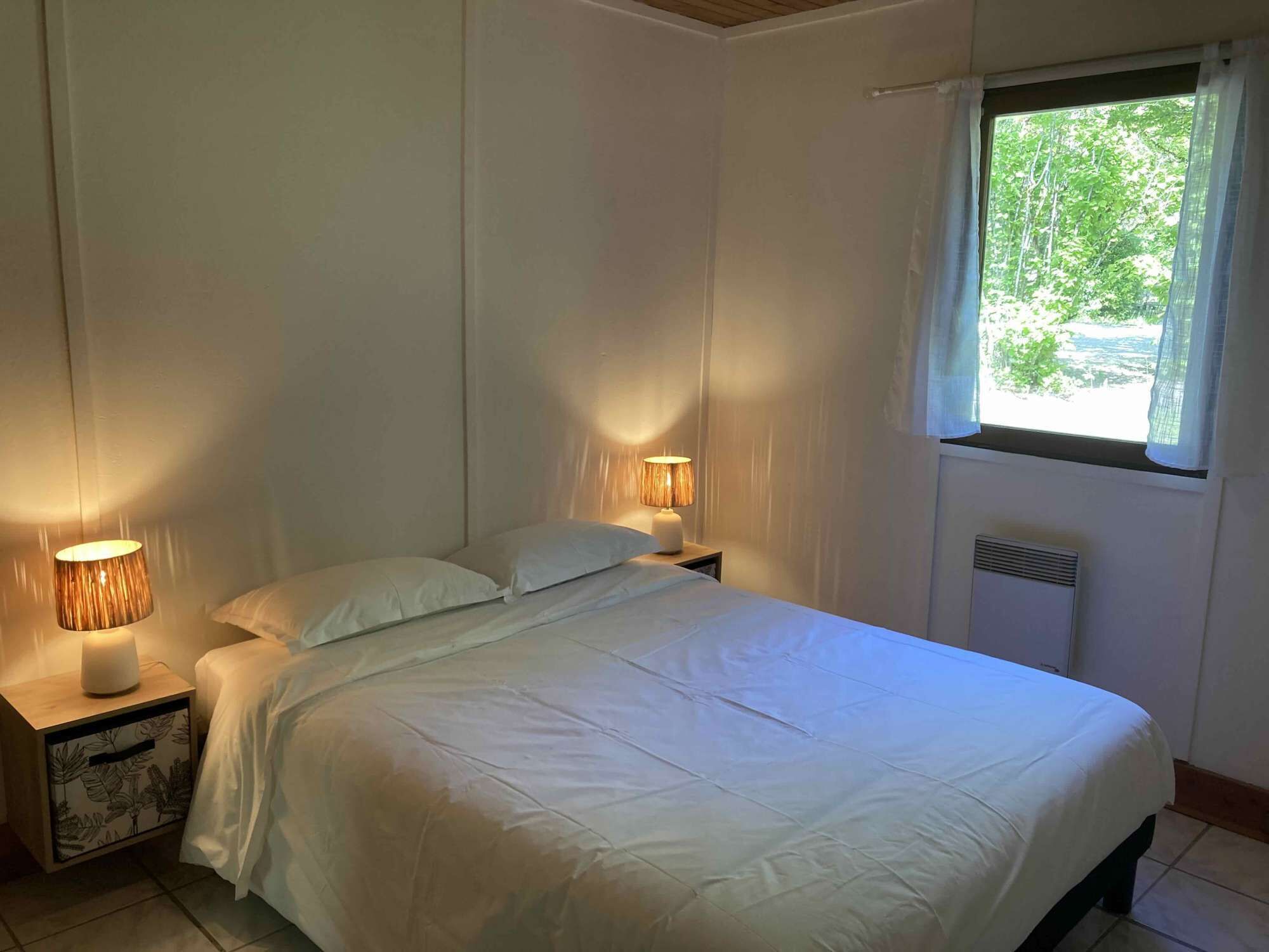 A bedroom with a bed, two bedside lamps, a window with curtains, and a radiator.