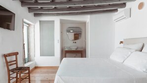A bedroom with a wooden floor, a bed with white linens, a wooden chair, a bedside table, and a mirror on the wall.