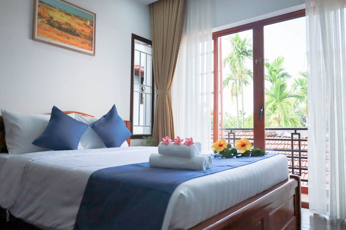 A neatly made bed with blue and white bedding, a window with a view of palm trees, and a painting on the wall.