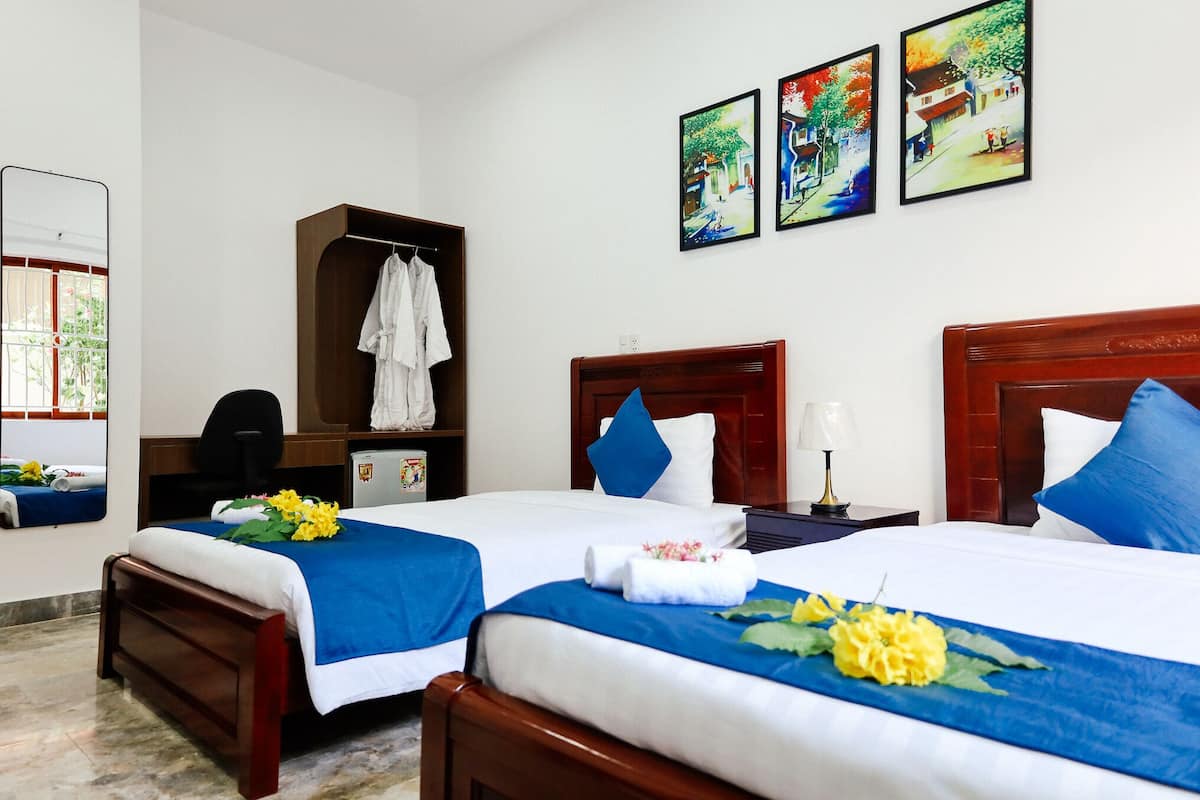 A hotel room with two beds, a wooden wardrobe, a desk with a chair, a mirror, and artwork on the wall.