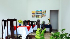 A dining area with wooden chairs and tables, some with red and white tablecloths. There are framed pictures on the wall and a small table with a vase of flowers. - Hoian Central Garden Homestay (Da Nang)