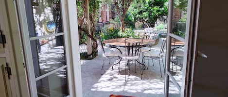 An open glass door leading to a patio with a round table and chairs, surrounded by greenery and a brick wall.