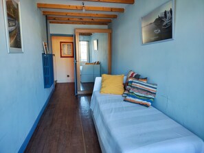 Ein schmaler Raum mit einem Bett, einer Holzbalkendecke und einer Tür zu einem weiteren Zimmer.