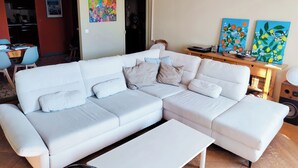 Un salón moderno con un sofá modular blanco, una mesa de centro blanca y una mesita de noche de madera con un altavoz. Hay dos cuadros en la pared, una cocina al fondo y una zona de comedor con sillas azules.