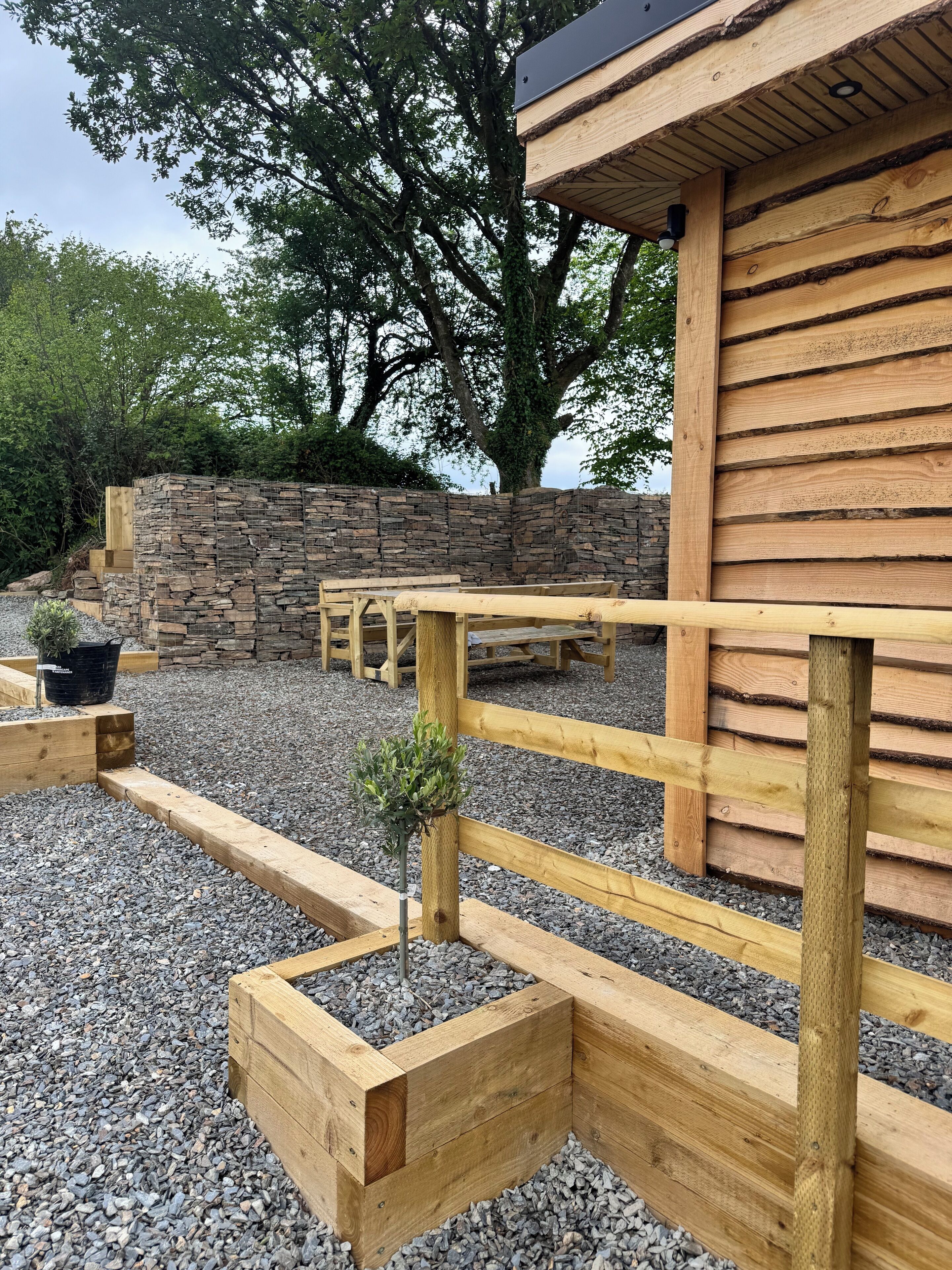 A wooden outdoor seating area with benches and planters, set against a stone wall and a building with a sloped roof.