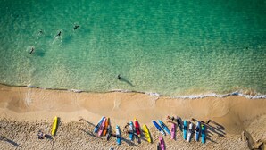 Vue aérienne d’une plage aux eaux turquoise et limpides, bordée d’un rivage sablonneux, où l’on aperçoit plusieurs kayaks colorés alignés sur le sable.