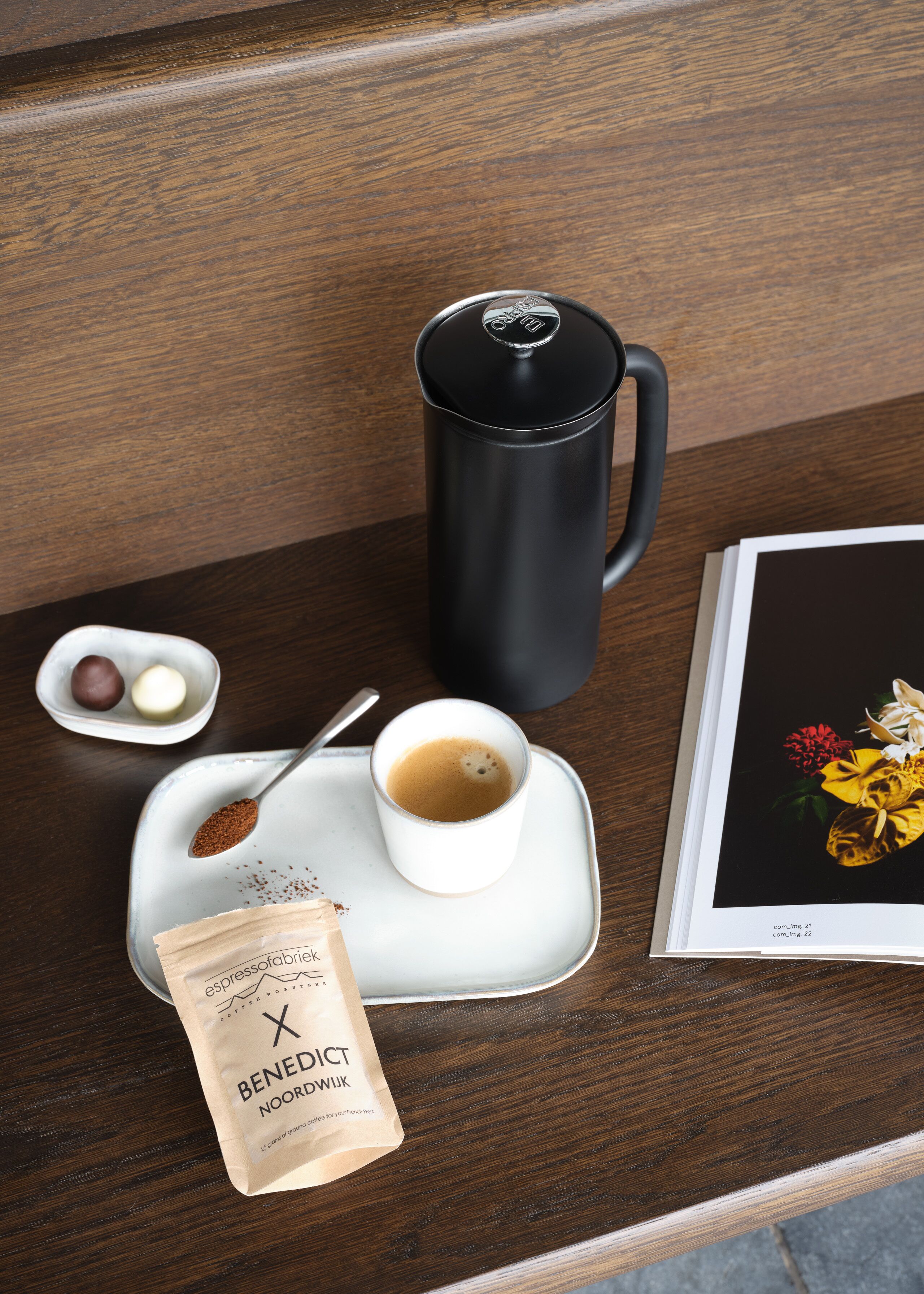 En kaffestation med en svart termos, en vit kaffekopp, en sked med kaffesump, en liten skål med två chokladbitar och en påse med kaffebönor märkta med texten ”Benedict Noordwijk”.