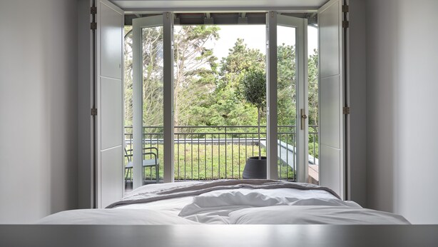 Un dormitorio con un ventanal grande, una cama y vistas a un balcón con una silla y vegetación.