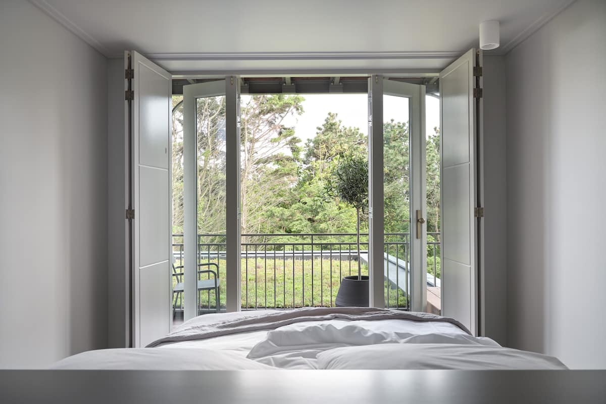 Ein Schlafzimmer mit großem Fenster, einem Bett und Blick auf einen Balkon mit Stuhl und Grünfläche.