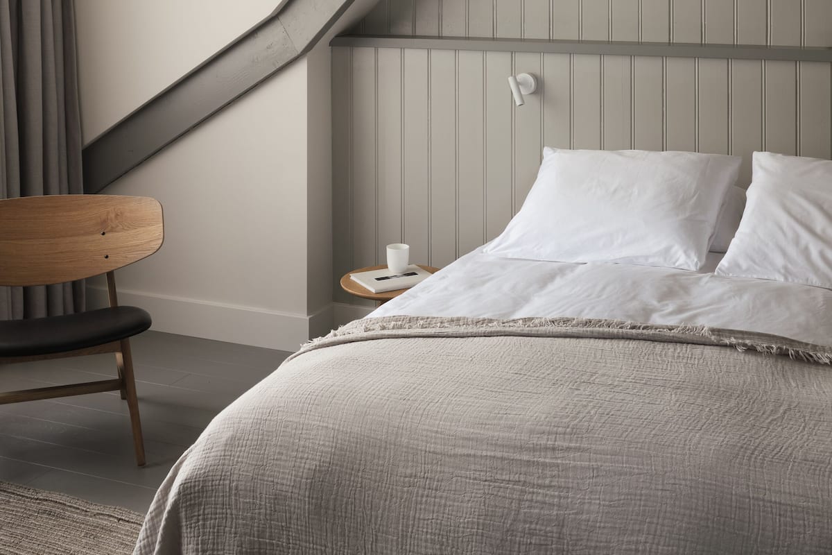 Ein Schlafzimmer mit schrägen Wänden, einem Holzstuhl, einem Nachttisch mit Tasse und einem Bett mit weißen Kissen.