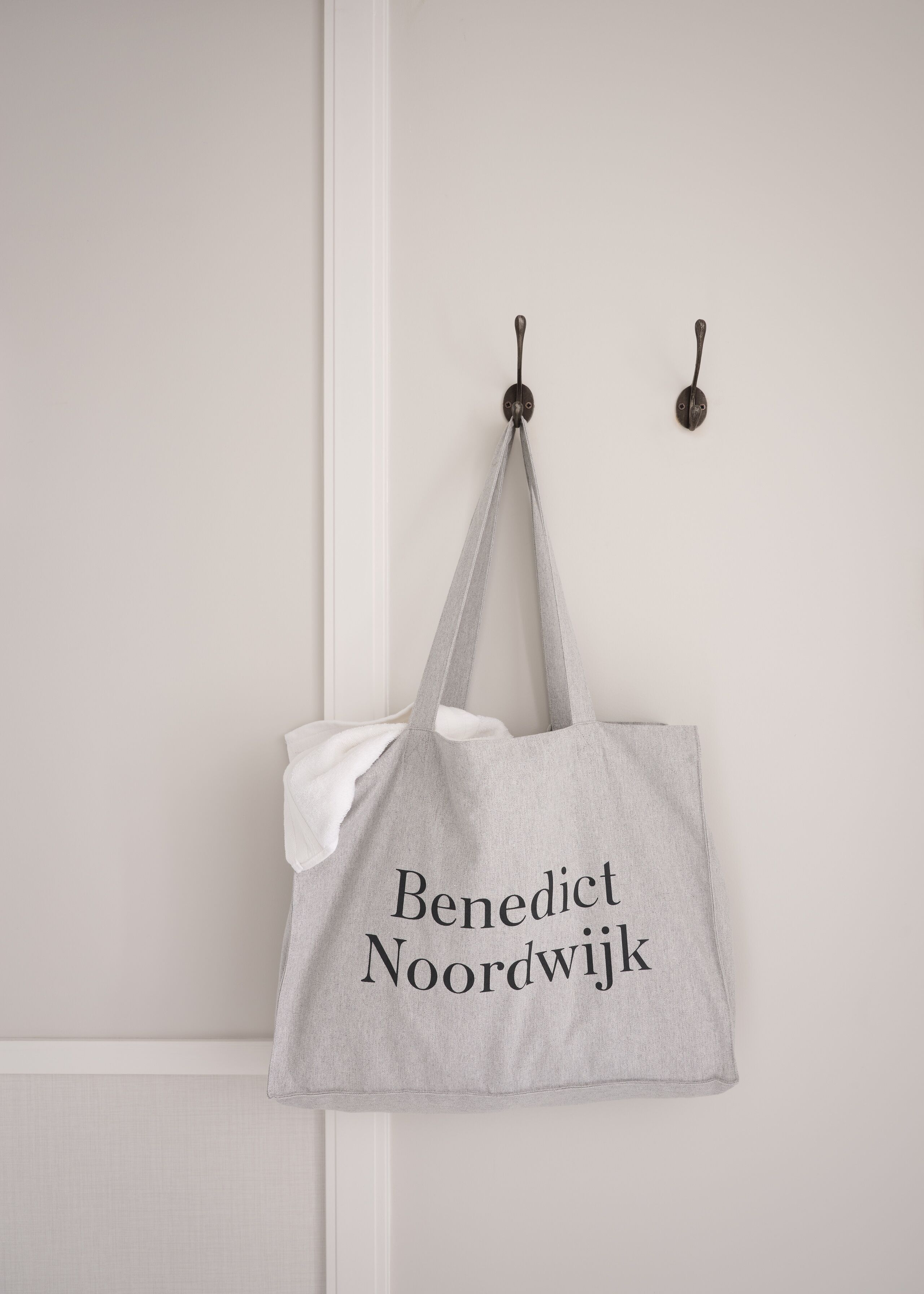 A grey tote bag with "Benedict Noordwijk" printed on it, hanging on a white door.