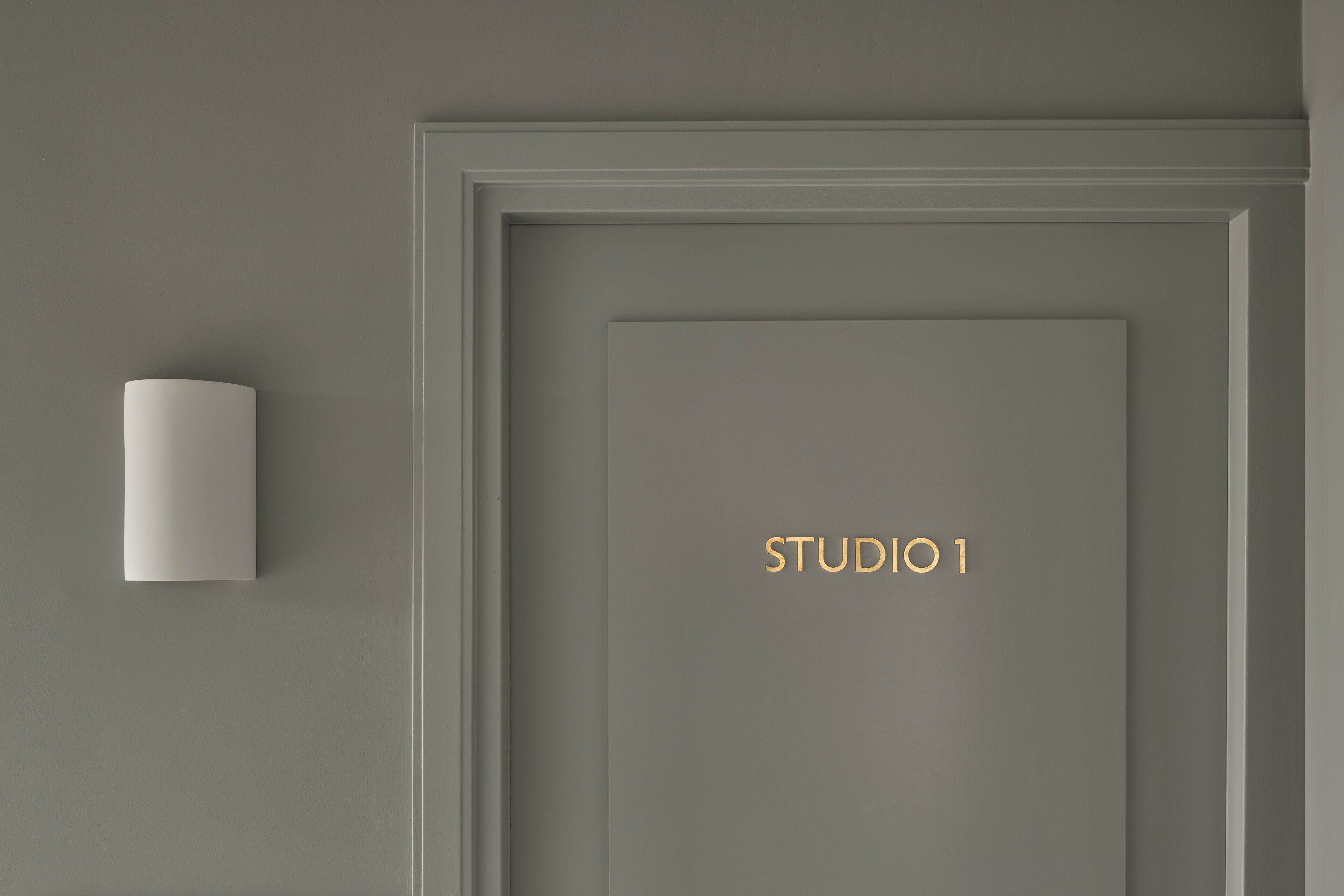 Eine minimalistische Tür mit der Aufschrift "STUDIO 1" in goldener Schrift, einer weißen Wandleuchte und einem schlichten, klaren Design.