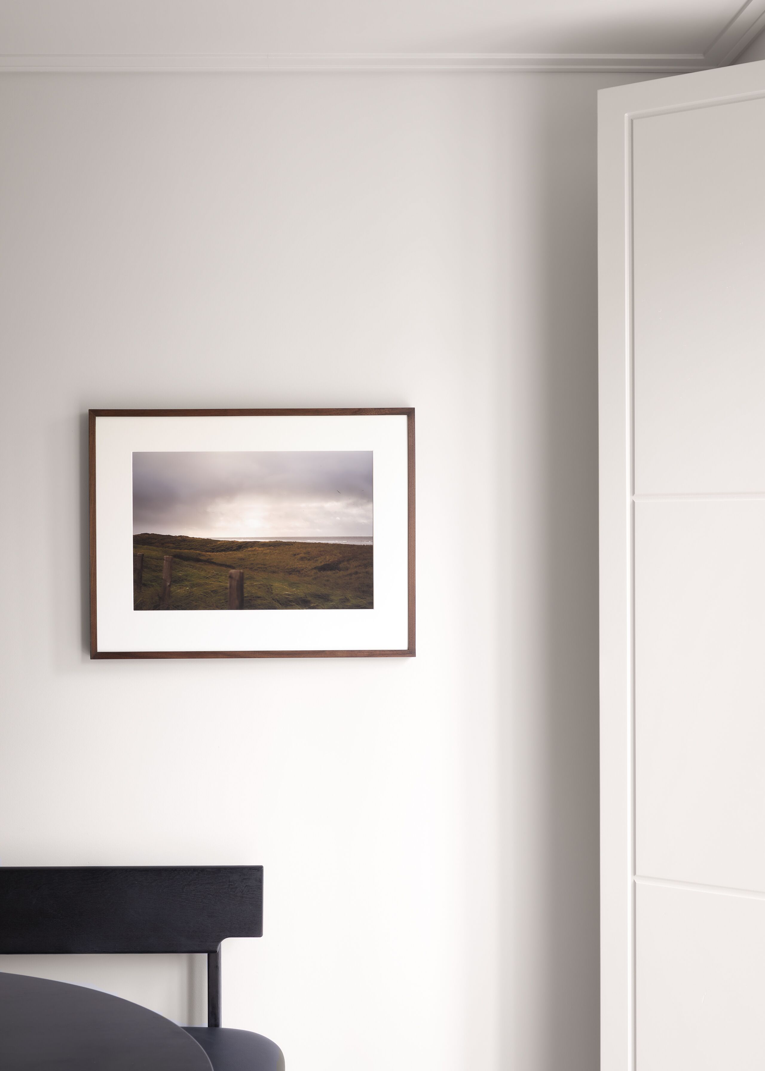 Une photographie de paysage encadrée sur un mur blanc, une chaise noire et une armoire blanche.