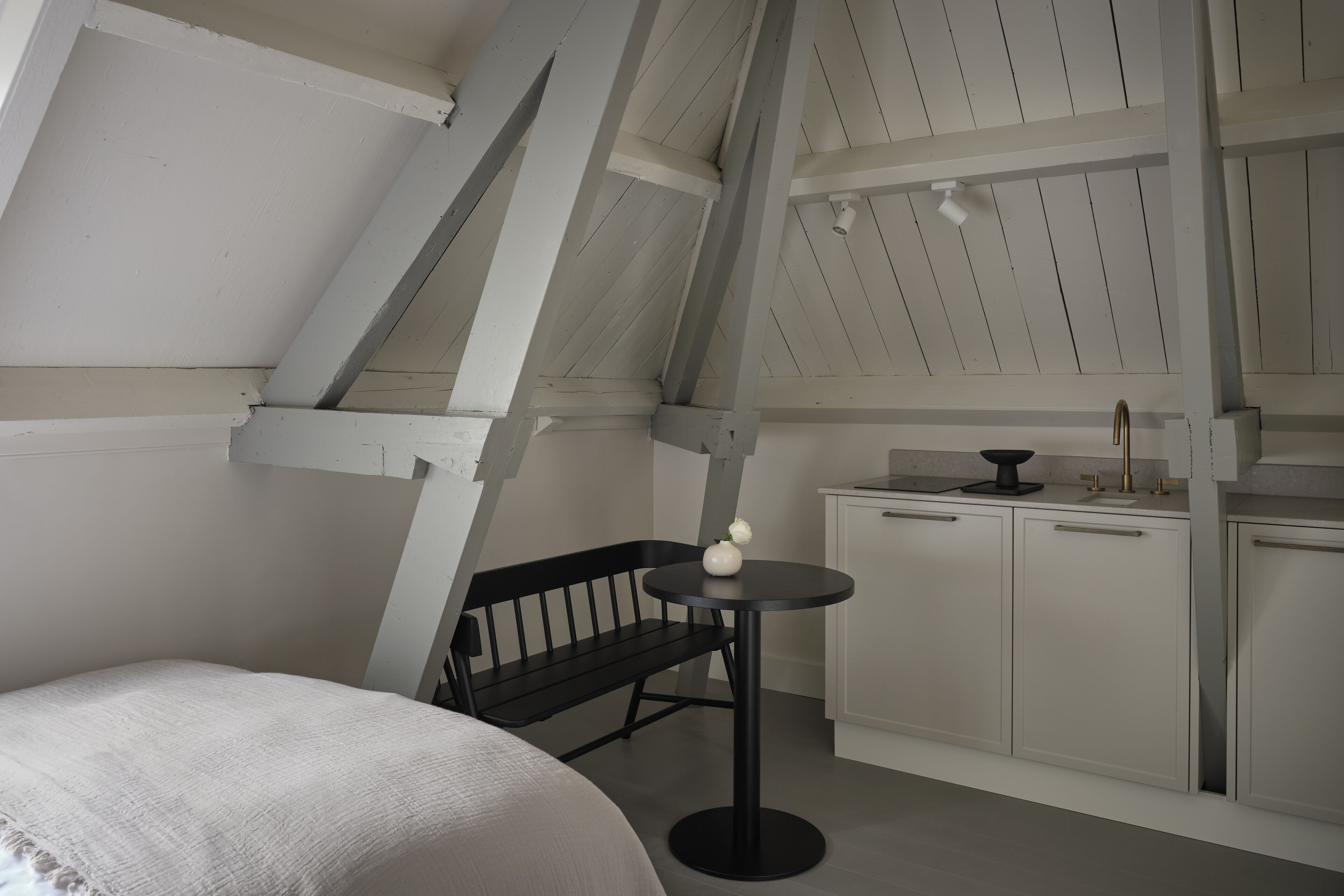 A room with a sloped ceiling, a black bench, a small round table with a vase, and white cabinets.