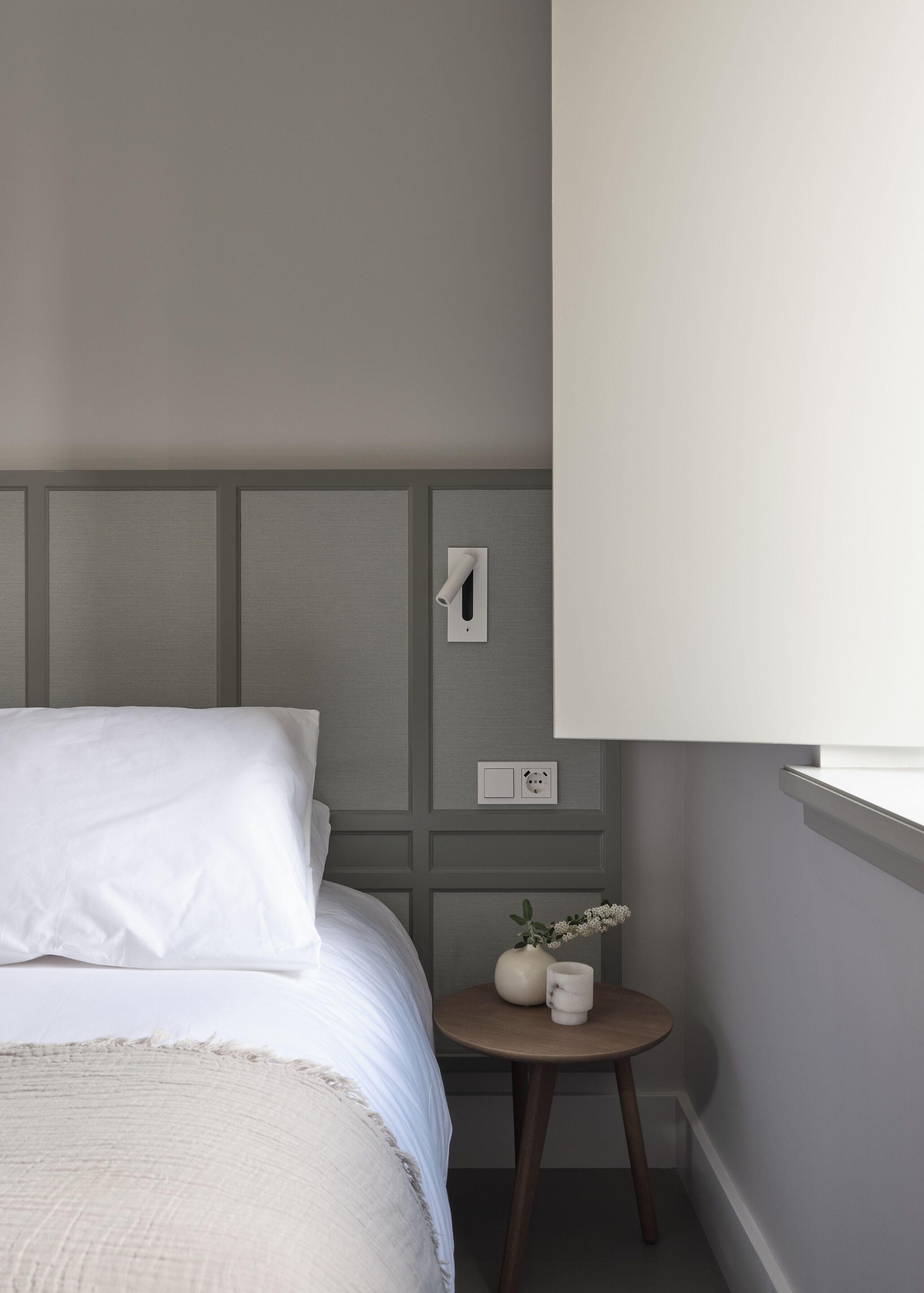 A bedroom with a bed, a small bedside table with a vase, and a wall socket.