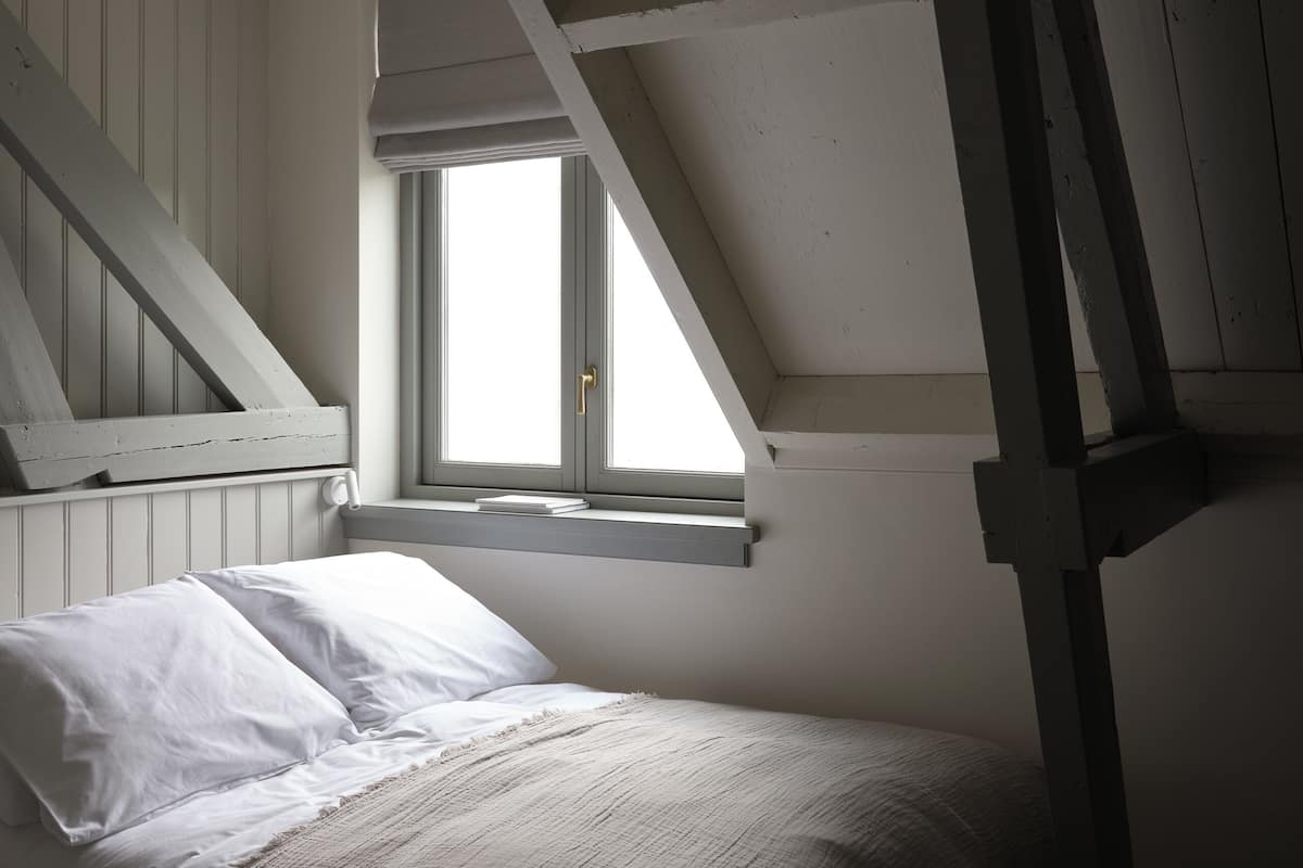 Ein Schlafzimmer mit schräg verlaufender Decke, einem Fenster, einem Bett mit weißer Bettwäsche und einer Holztreppe.