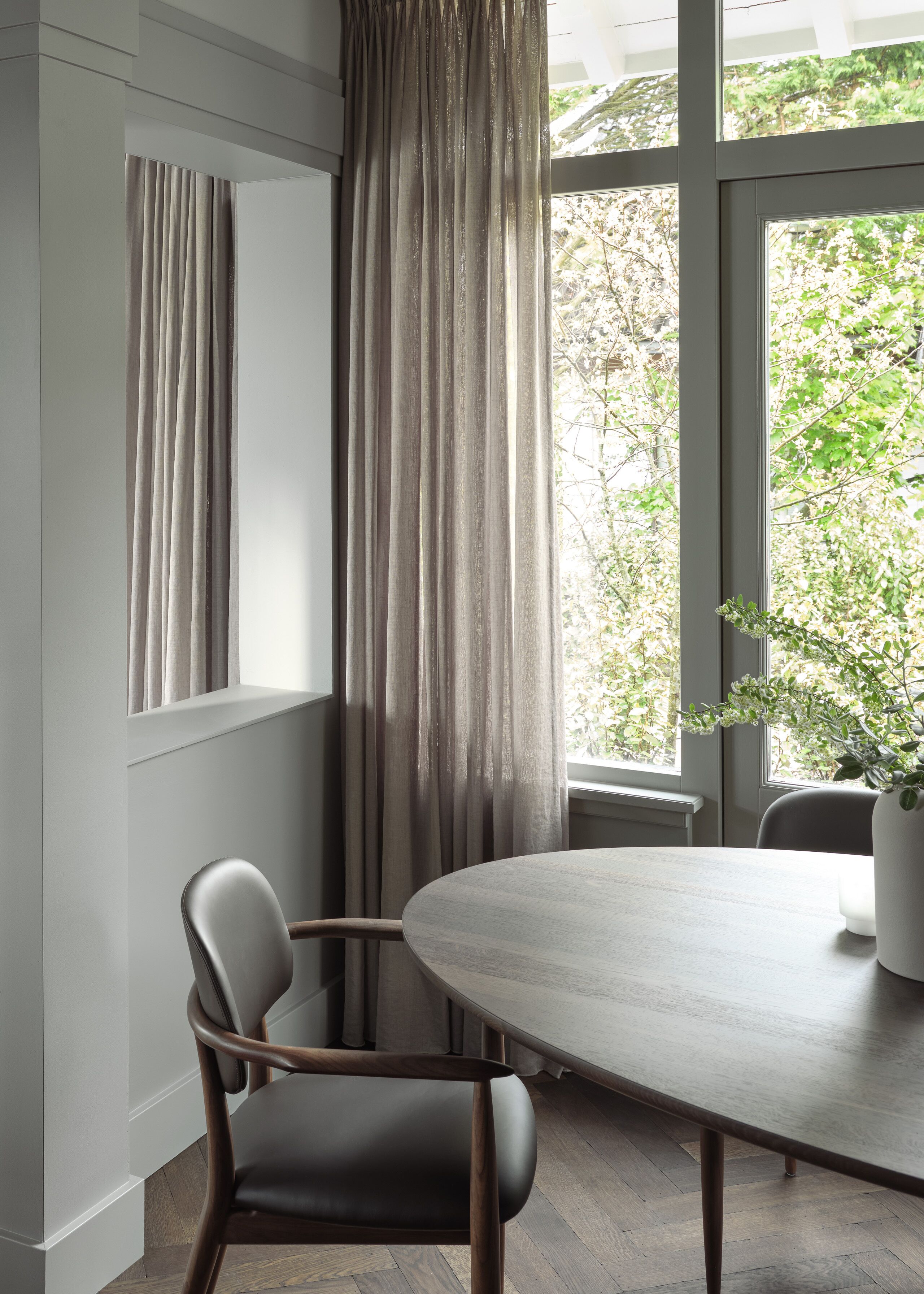Un comedor moderno con una mesa redonda y dos sillas, ventanas grandes con cortinas y una planta en el alféizar.