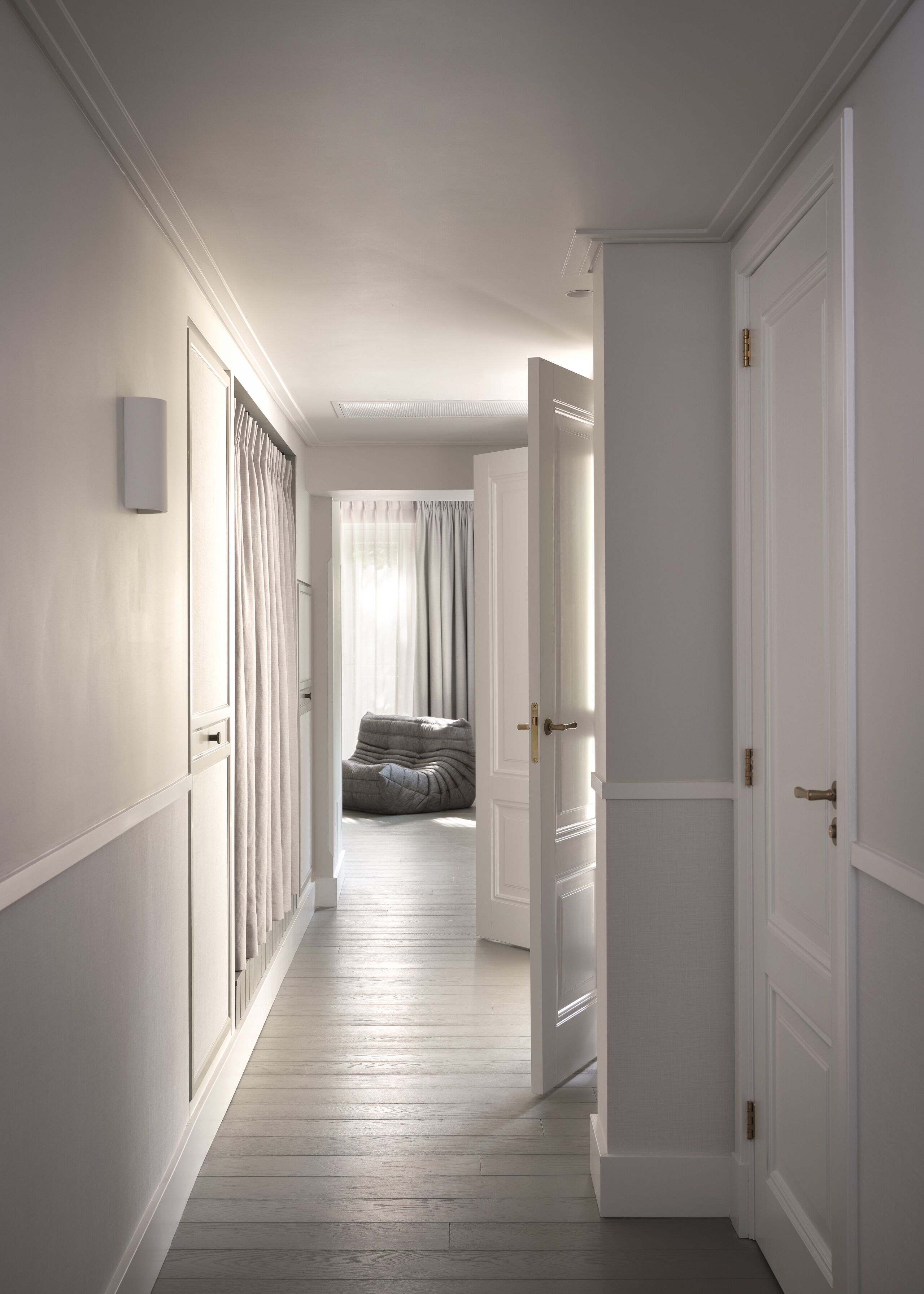 Un pasillo con paredes blancas, piso de madera y una puerta que da a una habitación con cama y cortinas.