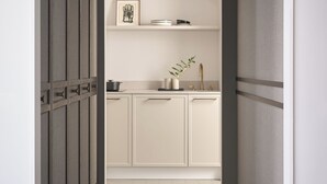 A narrow hallway leading to a kitchen with white cabinets, a sink, and a countertop with a plant and a framed picture. - Benedict Noordwijk (Noordwijk)