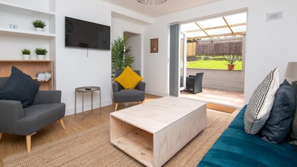 A modern living room with a sofa, coffee table, and TV. There is a small side table and a chair with a yellow pillow. The room has a large window and a view of the outside. - Newly Refurbished Gem | Perfect | Alexa | Sleeps 5 (Bristol)