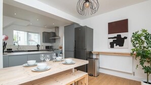 A modern kitchen with grey cabinets, a wooden dining table set with white dishes and glasses, a stainless steel trash bin, and a potted plant. - Newly Refurbished Gem | Perfect | Alexa | Sleeps 5 (Bristol)