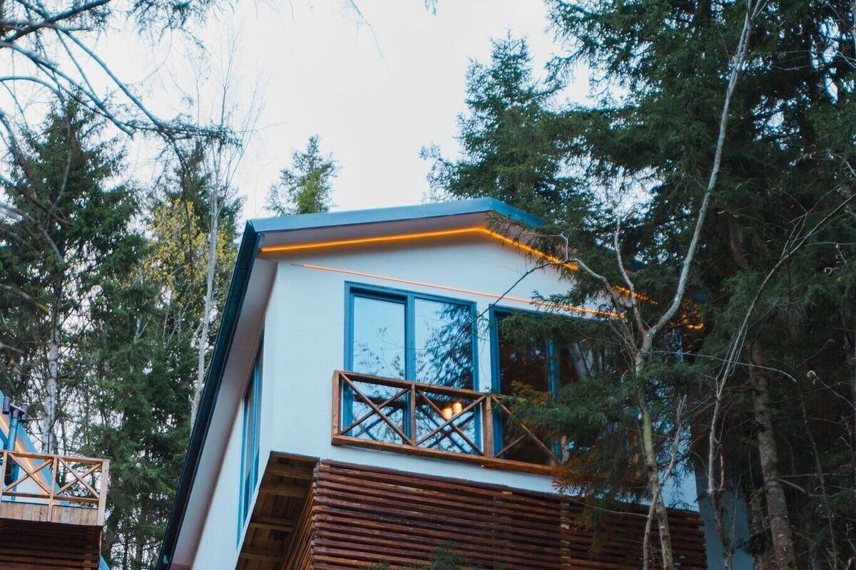 Una cabaña moderna con balcón, rodeada por un bosque frondoso.