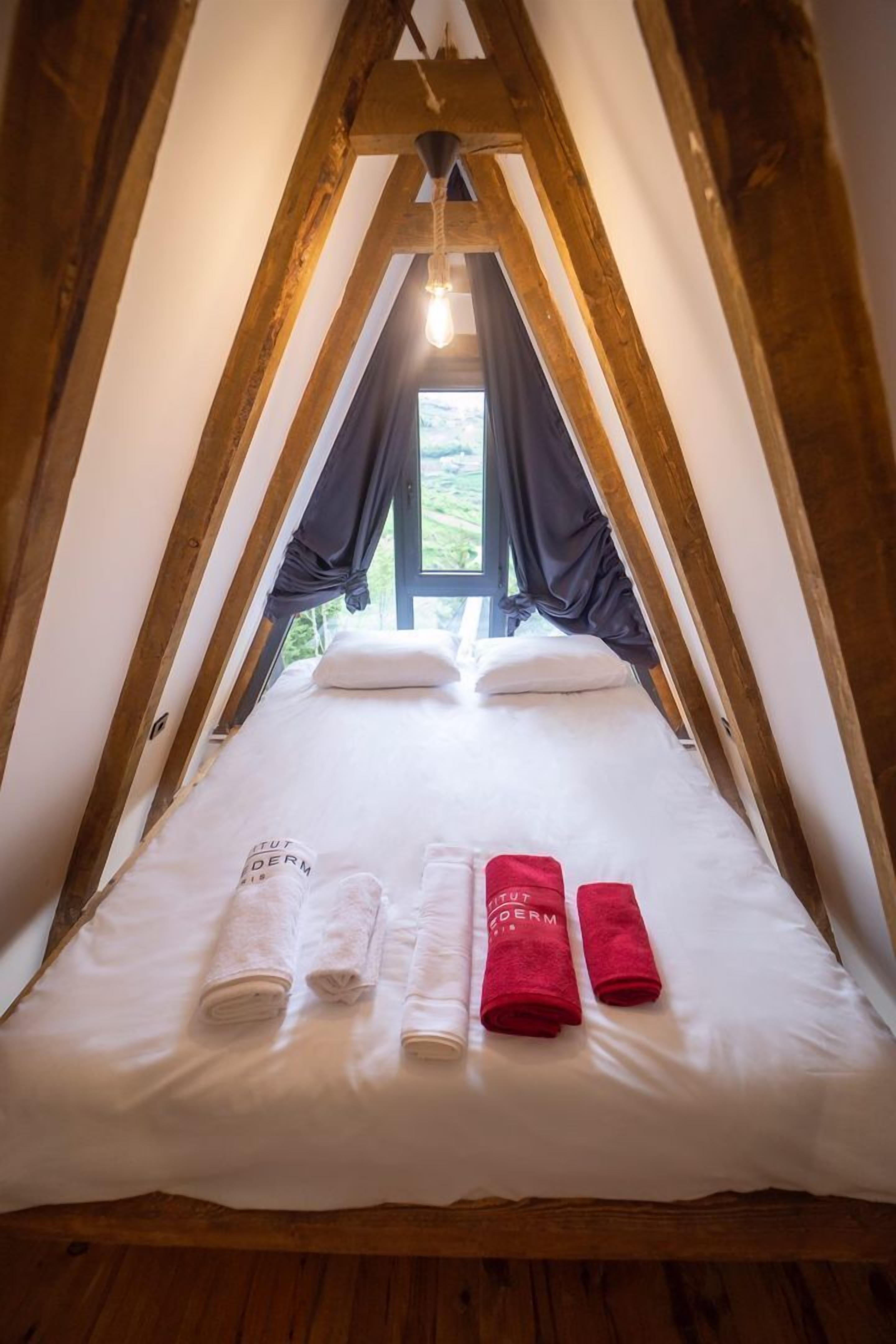 Una habitación con techo inclinado, una cama con ropa de cama y toallas blancas, y un ventanal con cortinas.