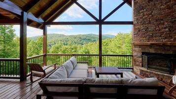 A covered outdoor seating area with a stone fireplace, overlooking a forested landscape.
