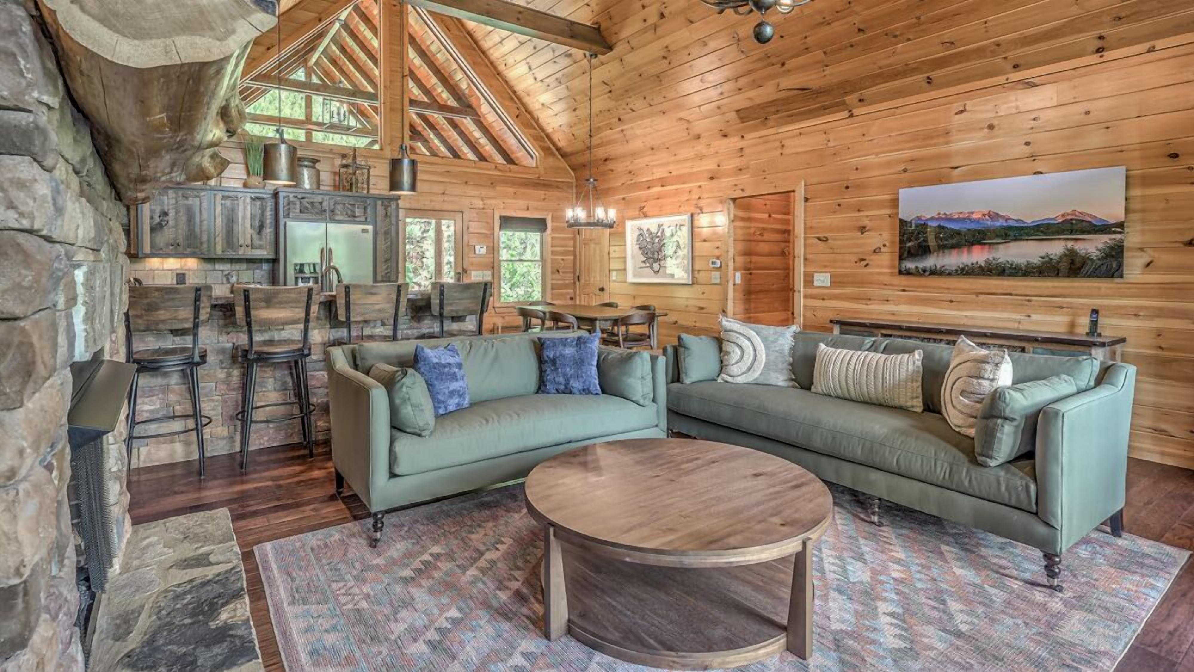 A cozy cabin interior with a stone fireplace, wooden dining area, and a large window overlooking a mountainous landscape.