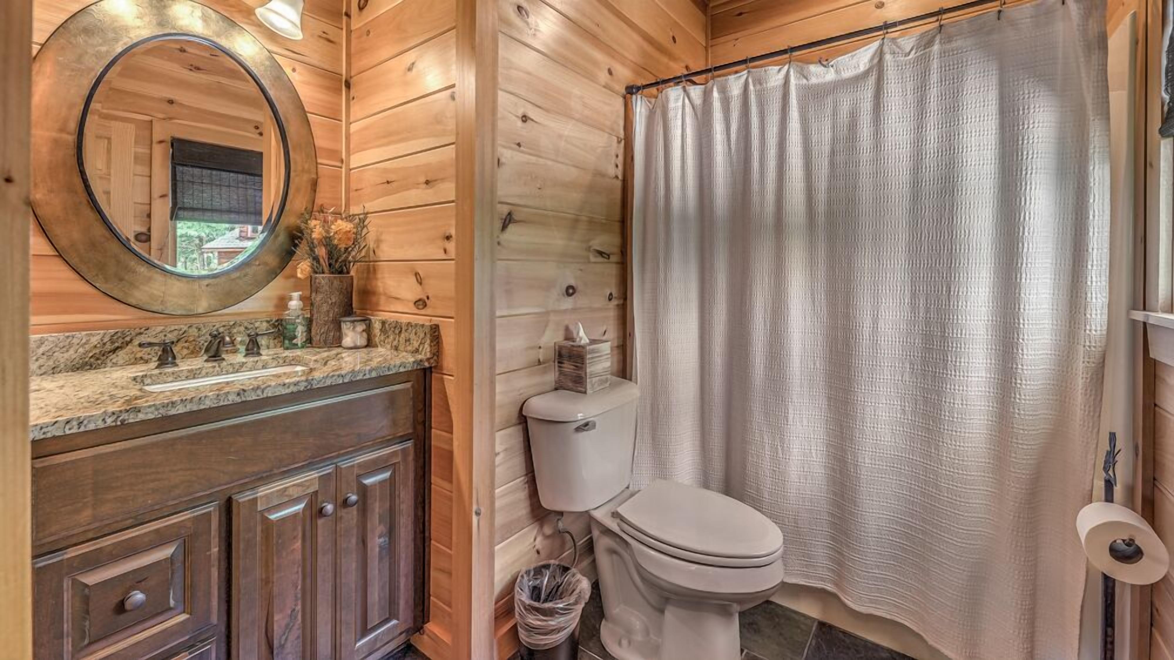 A wooden cabin bathroom with a round mirror, a sink with a marble countertop, a toilet, and a shower curtain.