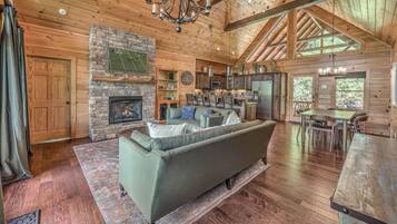 A cozy cabin interior with a stone fireplace, wooden walls, a dining area, and a kitchen.