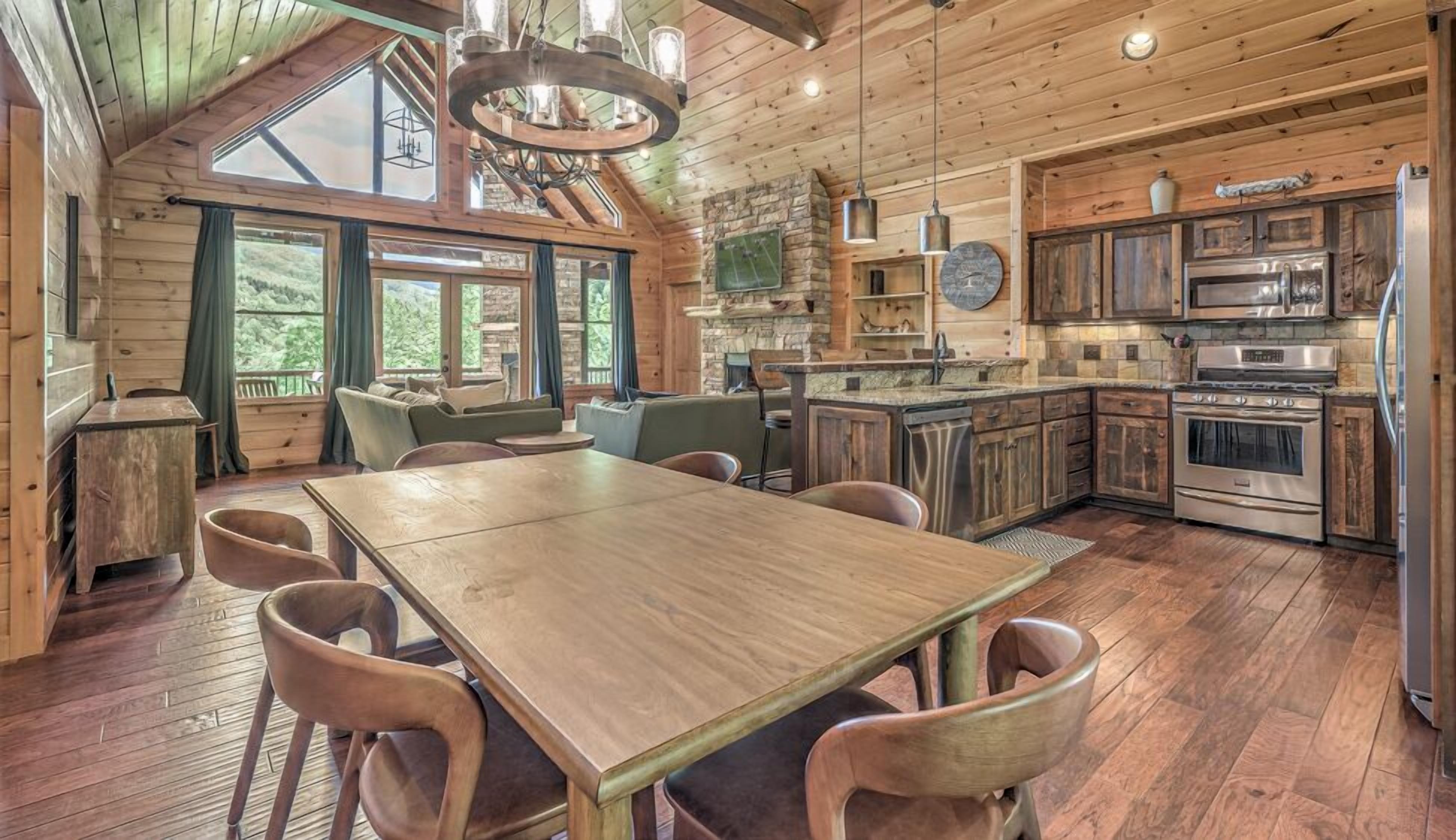 A rustic cabin interior with a wooden dining table, chairs, a stone fireplace, and a kitchen with modern appliances.